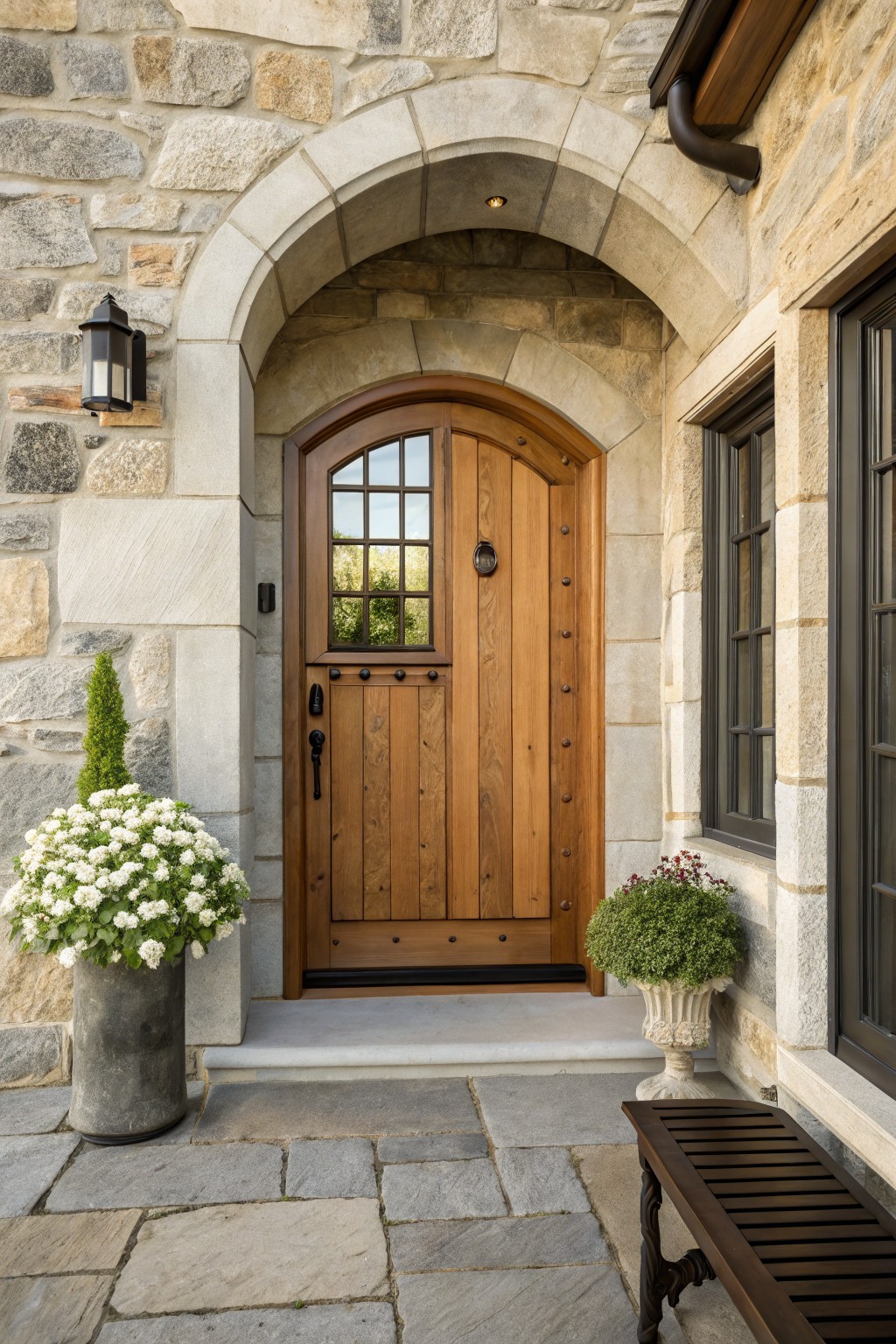 Arched wooden front door with glass panel and iron hardware set in a stone archway, flanked by potted plants and a bench on stone pavers.