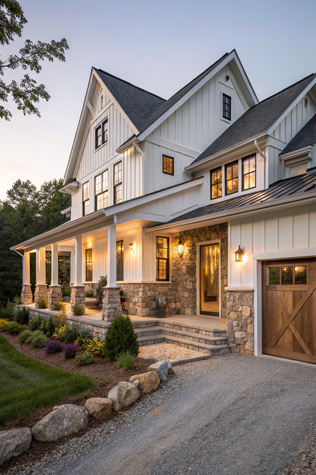 Two-story modern farmhouse exterior with white board-and-batten siding, gabled roof, wraparound porch supported by stone-based columns, stone entry surround and steps, wooden garage door, landscaping with plants and rock edging, and gravel driveway at dusk.