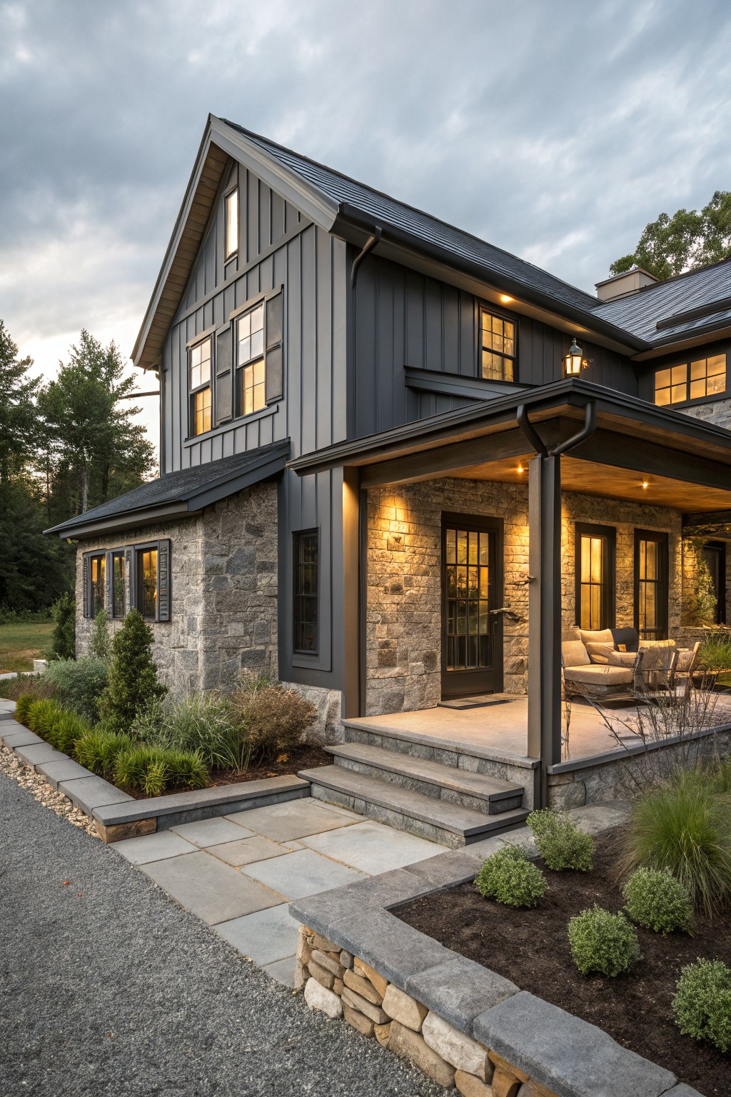 Corner view of a two-story modern farmhouse house with dark gray board-and-batten siding, gray stone foundation and lower walls, black-framed windows, covered front porch with furniture, stone steps, gravel driveway, and low plantings.