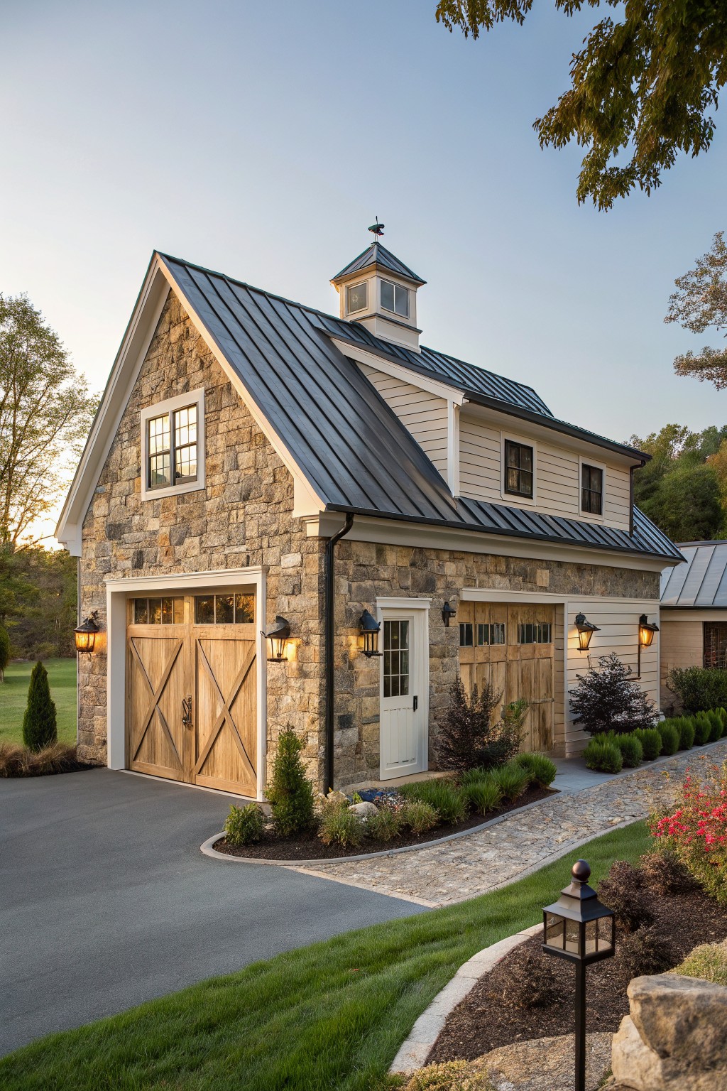 A detached two-story garage with fieldstone lower walls, board-and-batten siding above, wooden X-braced garage doors, black standing seam metal roof, cupola, and surrounding landscaping with driveway and path.