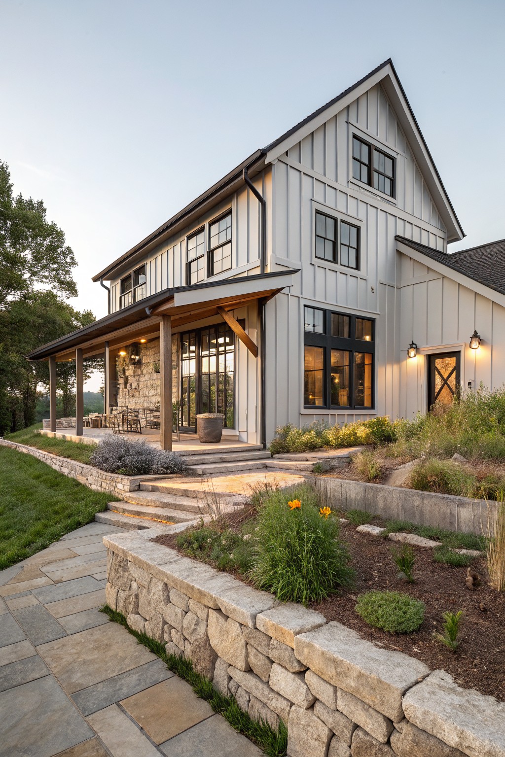 Two-story modern farmhouse exterior with white board-and-batten siding, covered porch supported by stone posts, black-framed windows and doors, stone retaining wall topped with plants and steps, paver walkway on grassy slope at dusk.