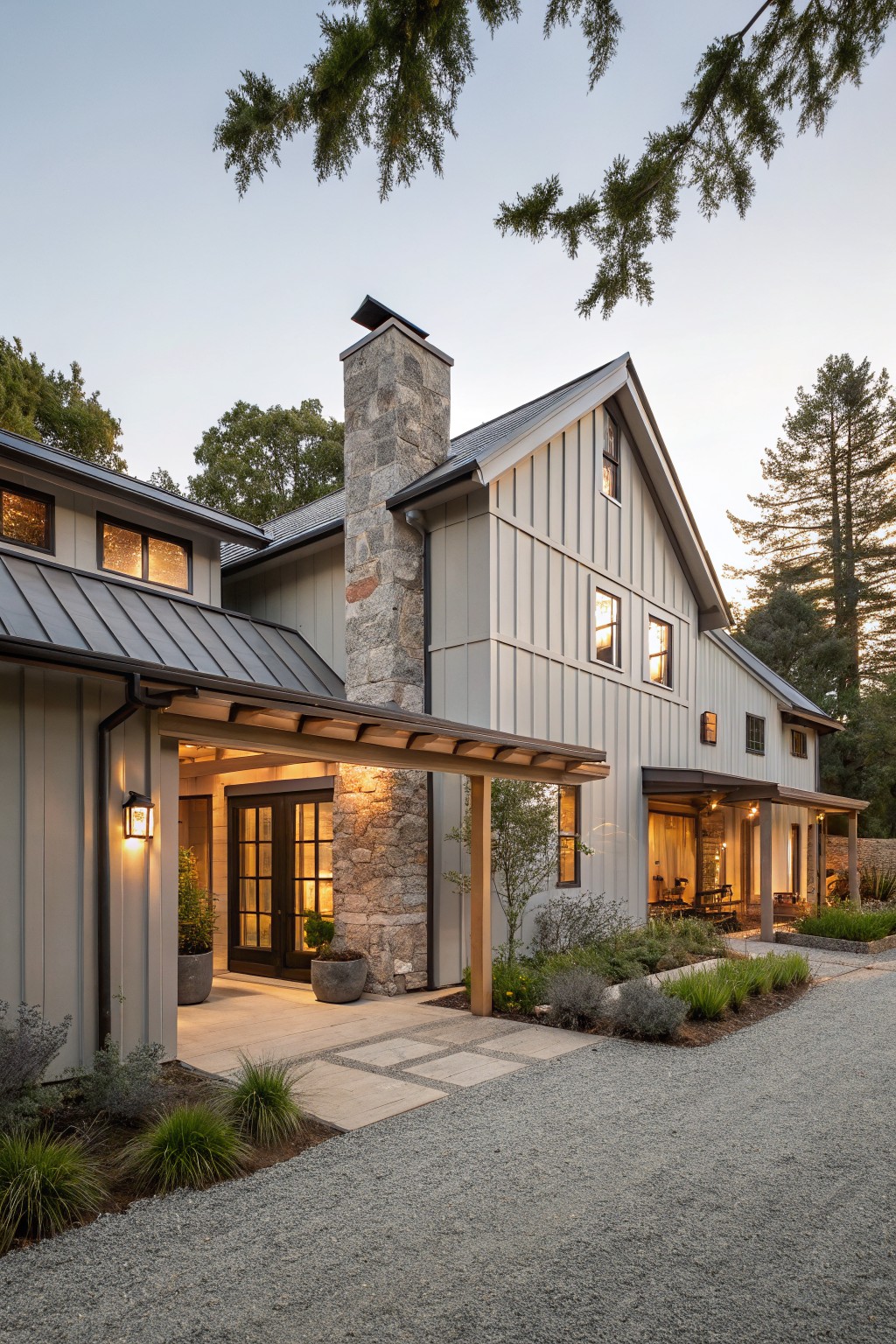 Modern farmhouse house exterior featuring board-and-batten siding, tall gray stone chimney, covered wooden porch with glass door entry, metal roof, surrounded by pine trees, plants, gravel driveway, and stone path at dusk.
