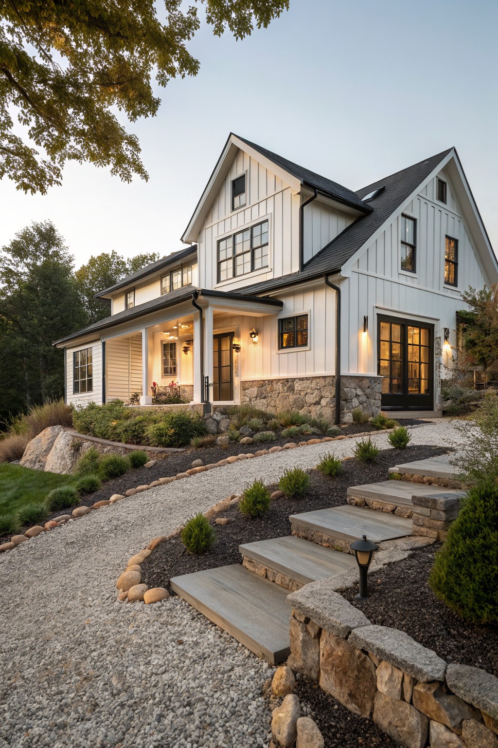 Two-story modern farmhouse exterior with white board-and-batten siding, black metal roof, large black-framed windows, wraparound porch with black door, fieldstone foundation, gravel driveway curving up stone steps amid boulders and shrubs on a wooded slope at dusk.