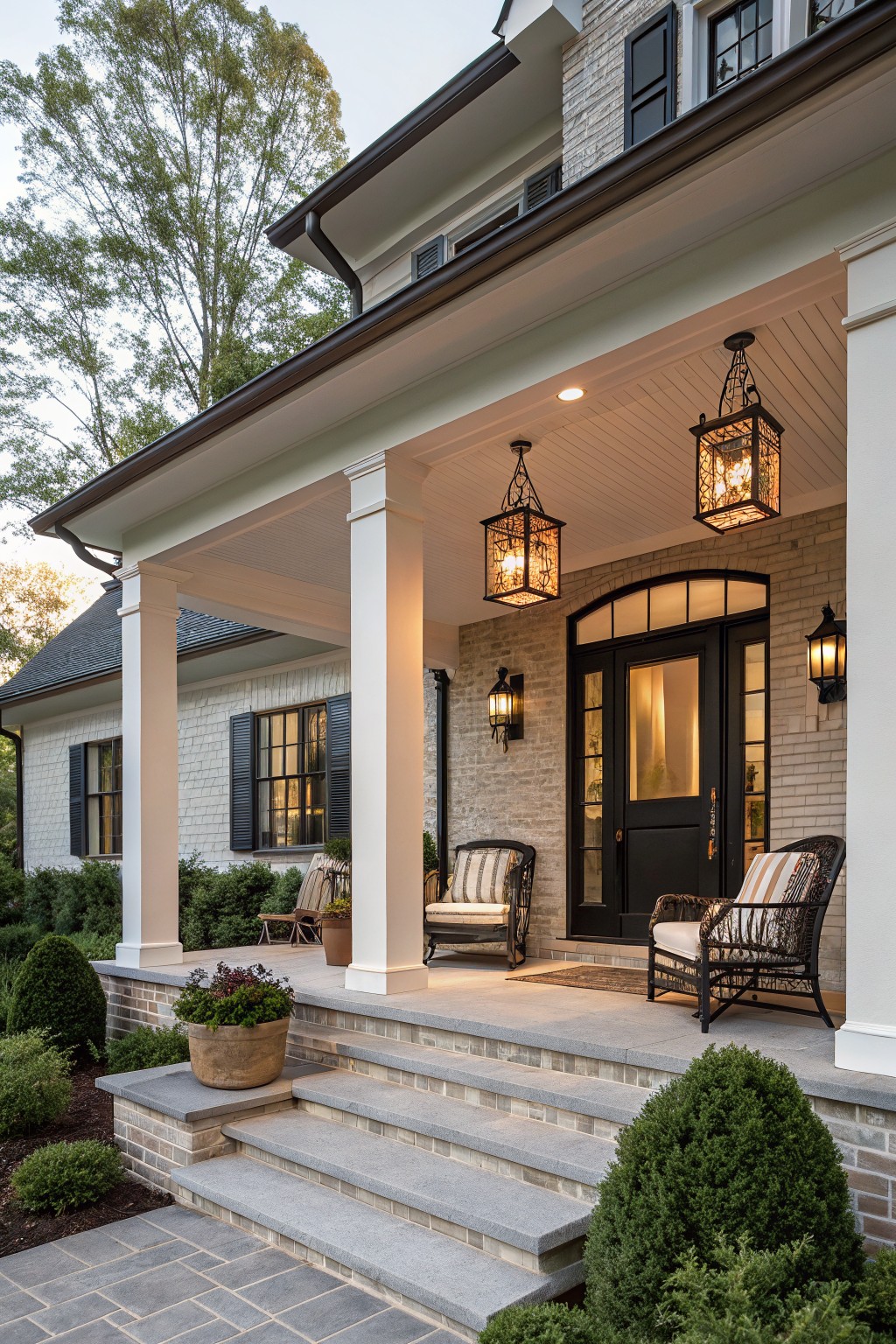 Front porch of a beige brick house with white columns, black double front doors, two hanging black lantern lights from the ceiling, wall sconces, wicker chairs with cushions, potted plants, stone steps, and surrounding shrubs and trees at dusk.