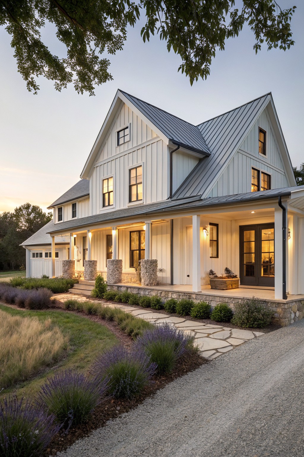 Two-story white board-and-batten farmhouse with dark metal roof, wraparound front porch on stone columns, garage wing, gravel driveway, and lavender borders at dusk.
