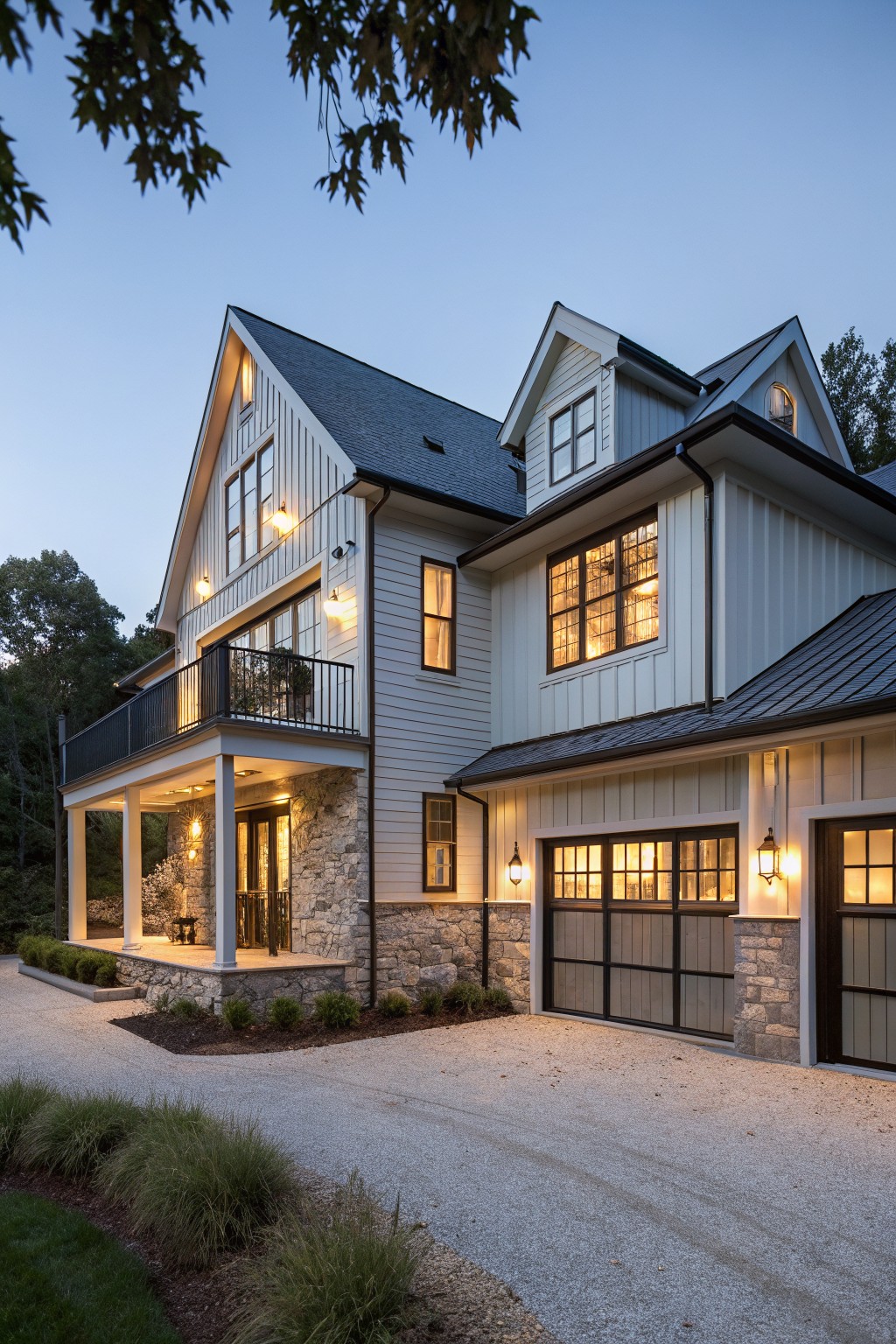 Two-story modern farmhouse house exterior at dusk with white board-and-batten siding, dark metal roof, stone base under covered front porch and garage doors, upper balcony, black metal railings, and gravel driveway edged by grasses and shrubs.
