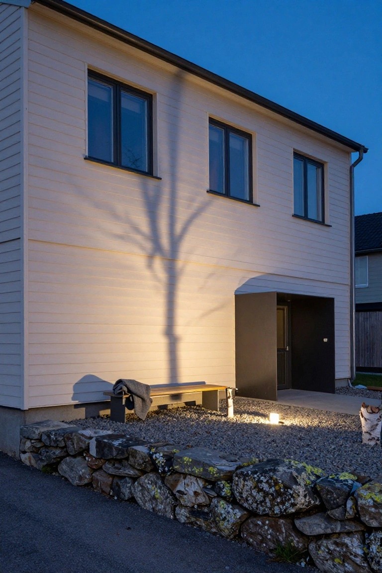 Modern white wood-clad house exterior at dusk illuminated by ground lights casting a large bare tree shadow on the wall, with dark-framed windows, recessed black entry door, wooden bench with blanket, stone retaining wall, gravel area, and nearby birch tree.
