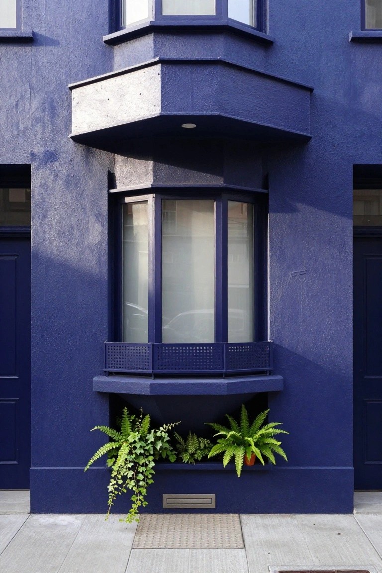 Deep blue painted house exterior with a curved bay window containing frosted glass panes, flanked by dark blue doors, ferns in a built-in planter below the window, and a concrete sidewalk in front.