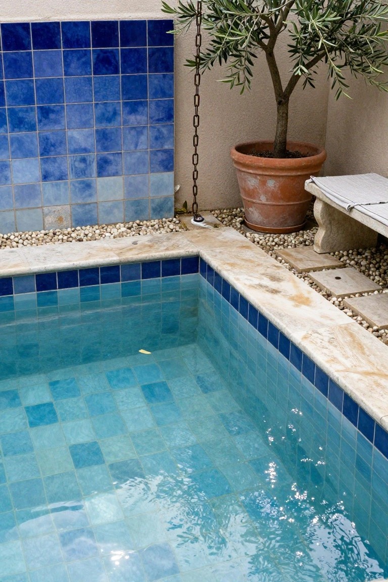 Corner swimming pool lined with gradient blue mosaic tiles on walls and bottom, adjacent stone bench, potted olive tree in terracotta pot, pebble mulch, and tiled backsplash wall.