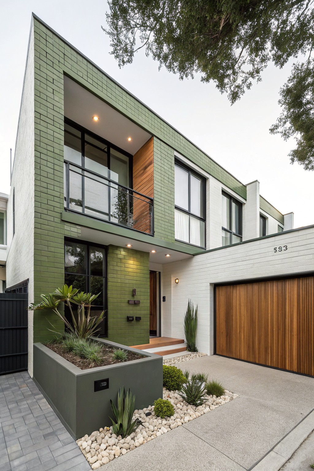 Modern two-story house exterior with green painted brick on angular walls, white rendered sections, timber garage door, glass balcony with black railing, and simple succulent landscaping in the front yard.