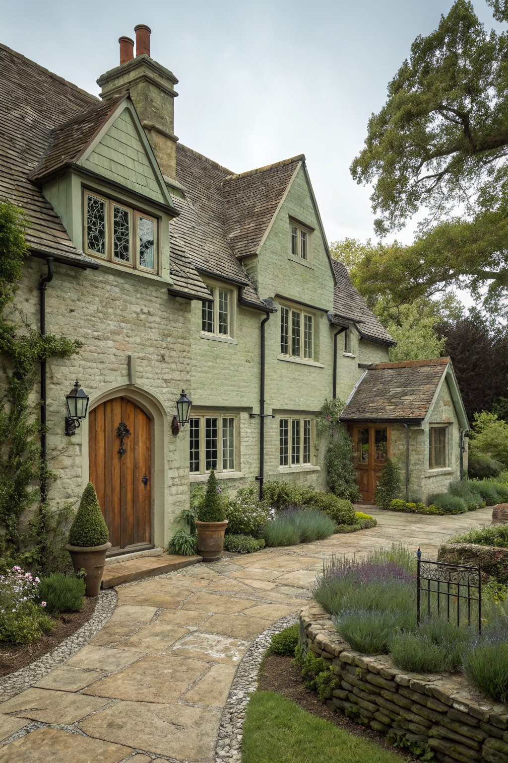 Two-story house with pale green painted stone walls, gabled roofs, arched wooden front door with lanterns, stone pathway, lavender plants, and surrounding greenery.