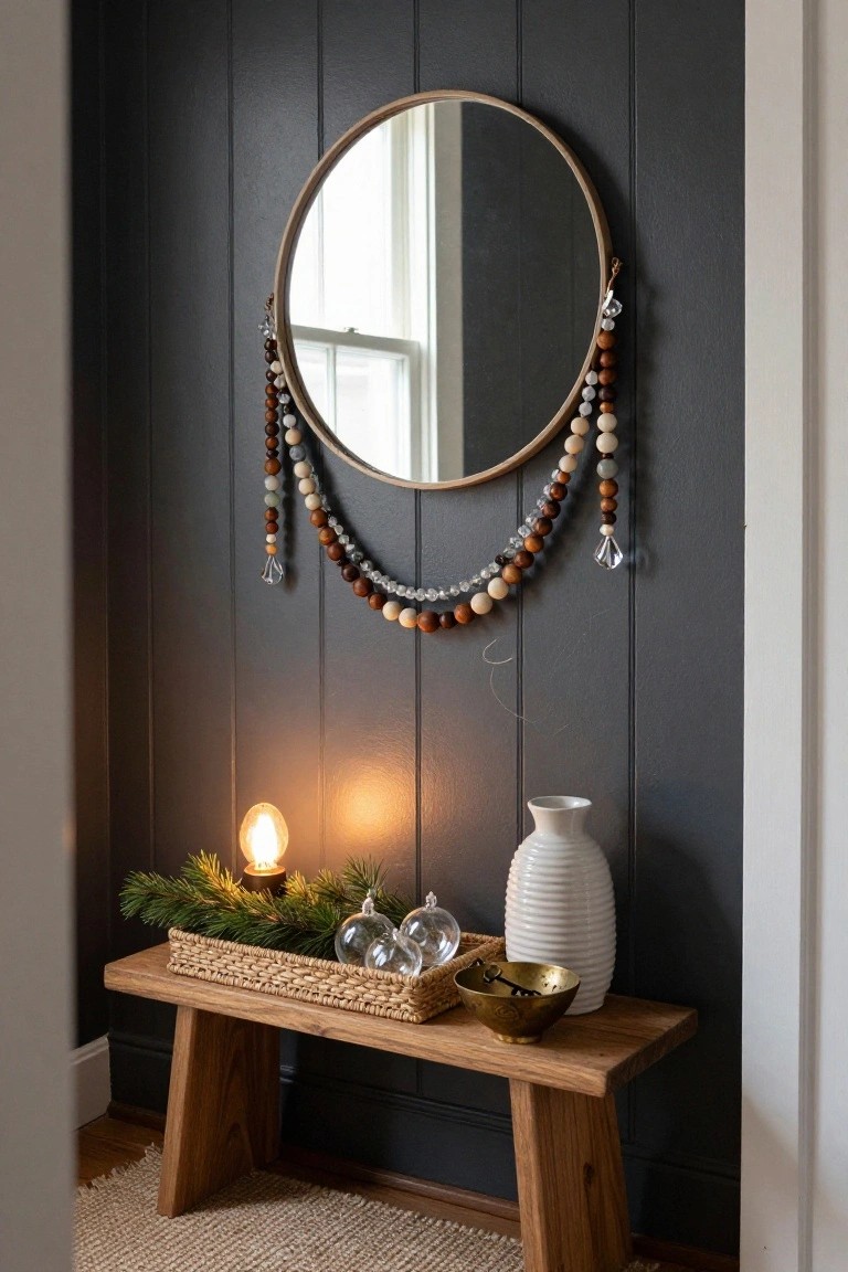 Dark wood-paneled wall with large round mirror hung with multicolored wooden and glass beaded garlands, above wooden bench holding wicker basket of pine branches and glass ornaments, lit candle, white vase, and brass bowl with keys.
