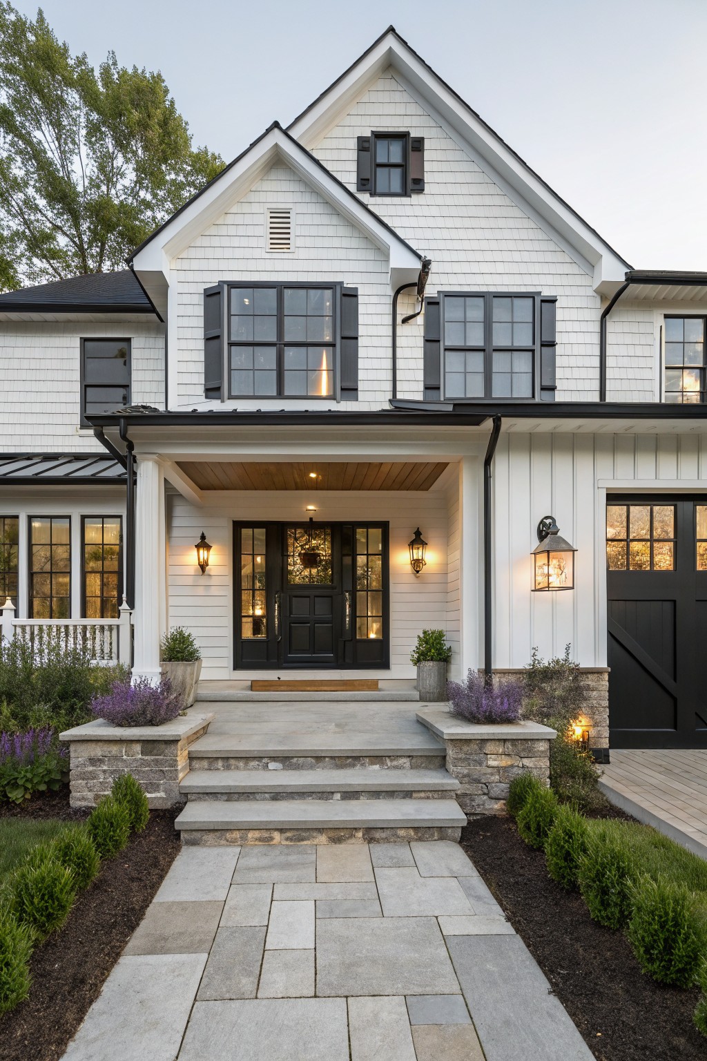 Front view of a two-story white shiplap-sided modern farmhouse with black-framed windows, shutters, front door, and trim, a covered wooden porch, stone steps, potted plants, and a flagstone path bordered by shrubs.