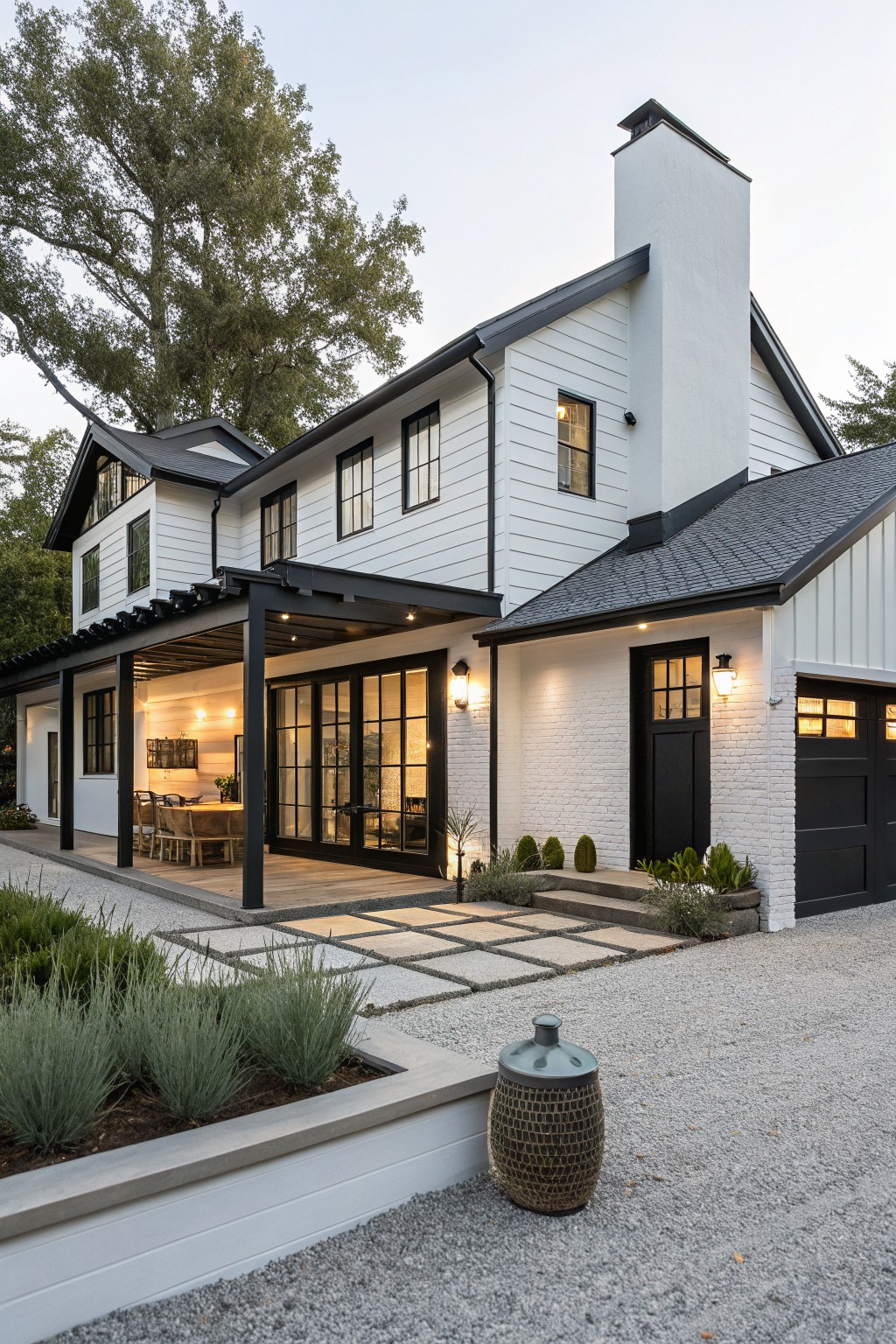 White board-and-batten sided modern farmhouse house exterior with black metal pergola over a patio dining area, black windows and doors, black garage, gravel driveway, and low landscaping.