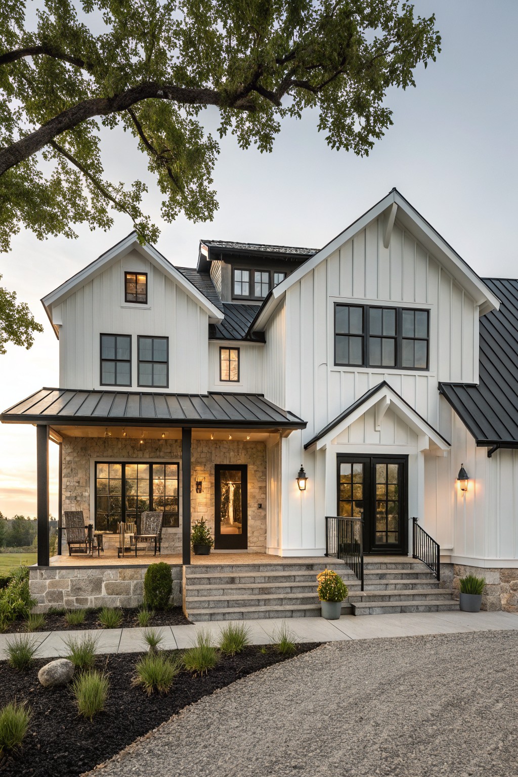 White board-and-batten farmhouse with black metal roof and gable dormers, covered front porch with stone base and black doors, steps leading to gravel driveway surrounded by low plantings at dusk.