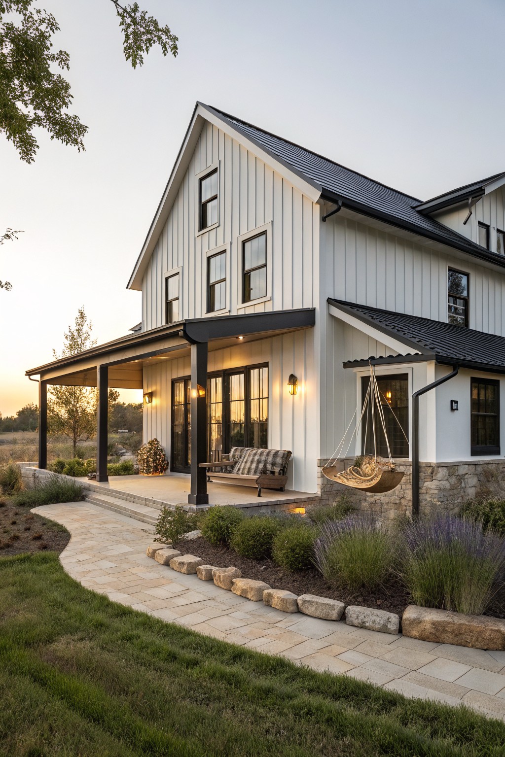 White board-and-batten sided modern farmhouse with black metal roof and trim, covered porch featuring a swing bed and bench, stone pathway, and low plantings around the base at dusk.