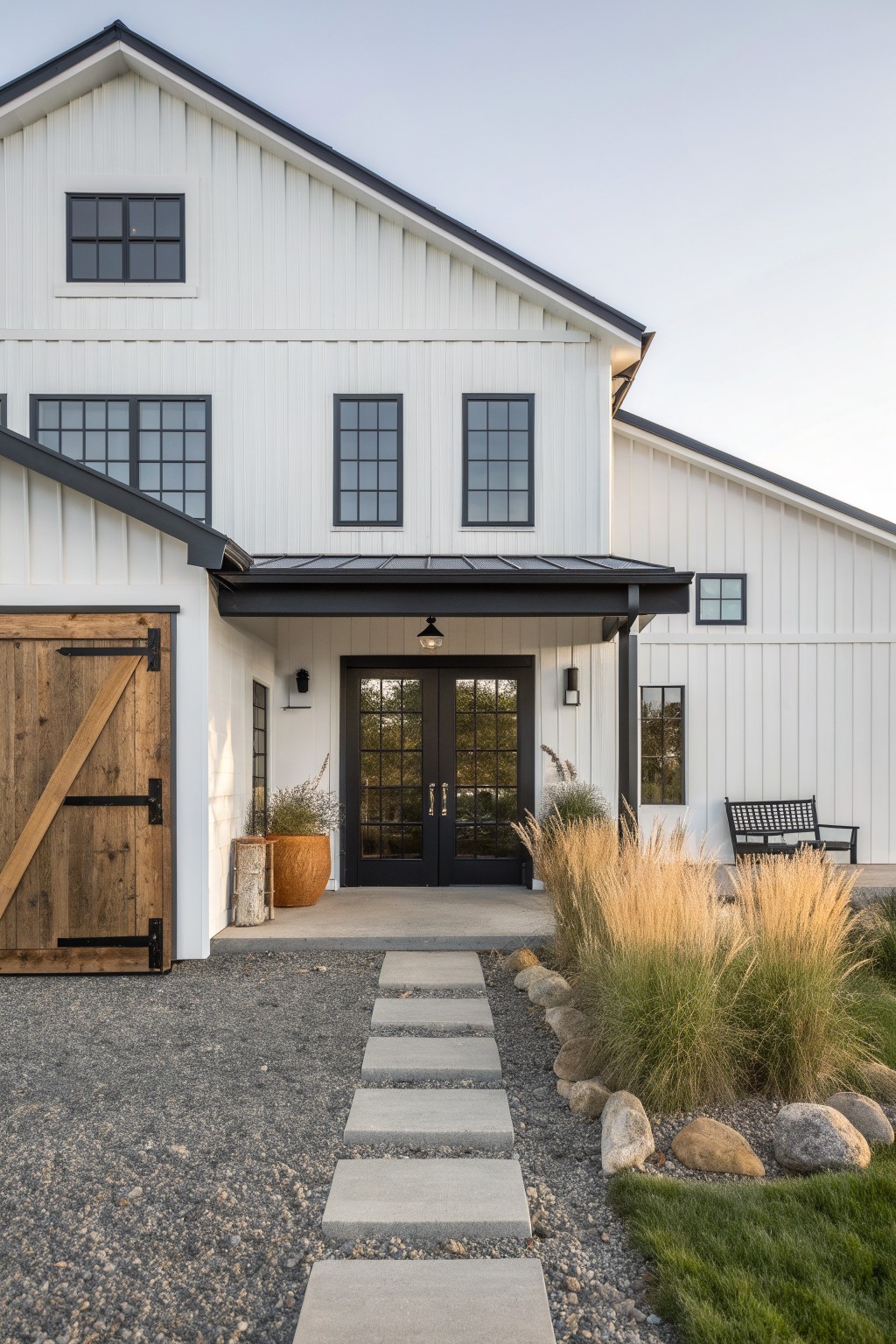 Two-story white board-and-batten sided modern farmhouse exterior with black metal roof overhangs, black-framed windows, wooden sliding barn-style garage door, black double entry doors under a covered porch, concrete paver pathway through gravel yard edged with grasses and rocks.