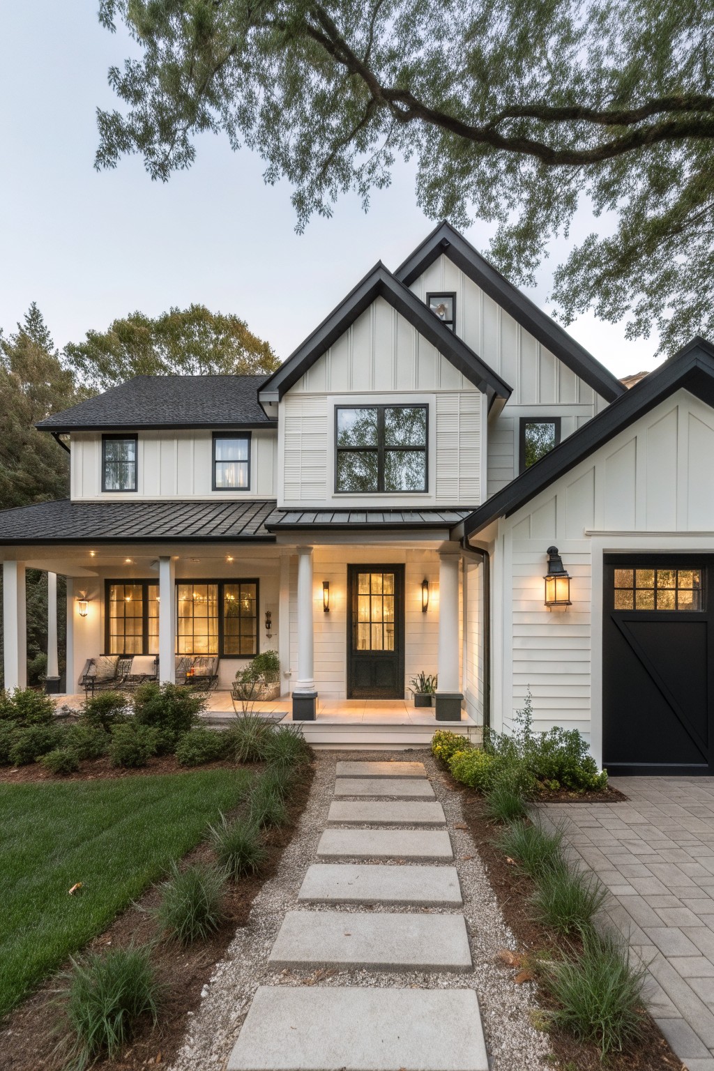 Two-story modern farmhouse exterior with white board-and-batten siding, black standing-seam metal roof, black-framed windows and trim, covered front porch with white columns and black door, attached garage, stone pathway, gravel edging, ornamental grasses, shrubs, and lawn under large tree branches at dusk.