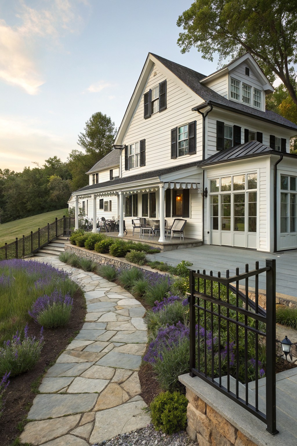 White clapboard farmhouse exterior with black metal roof, shutters, wraparound porch, black railings, and a flagstone path lined with lavender plants leading to a deck.