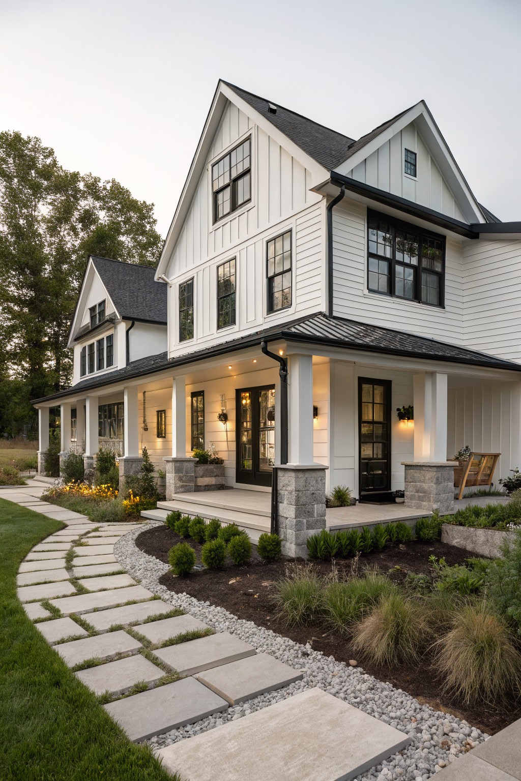 White board-and-batten sided modern farmhouse exterior with black metal roof, black-framed windows and doors, covered front porch supported by stone pillars, and a curved stone pathway through landscaped front yard.