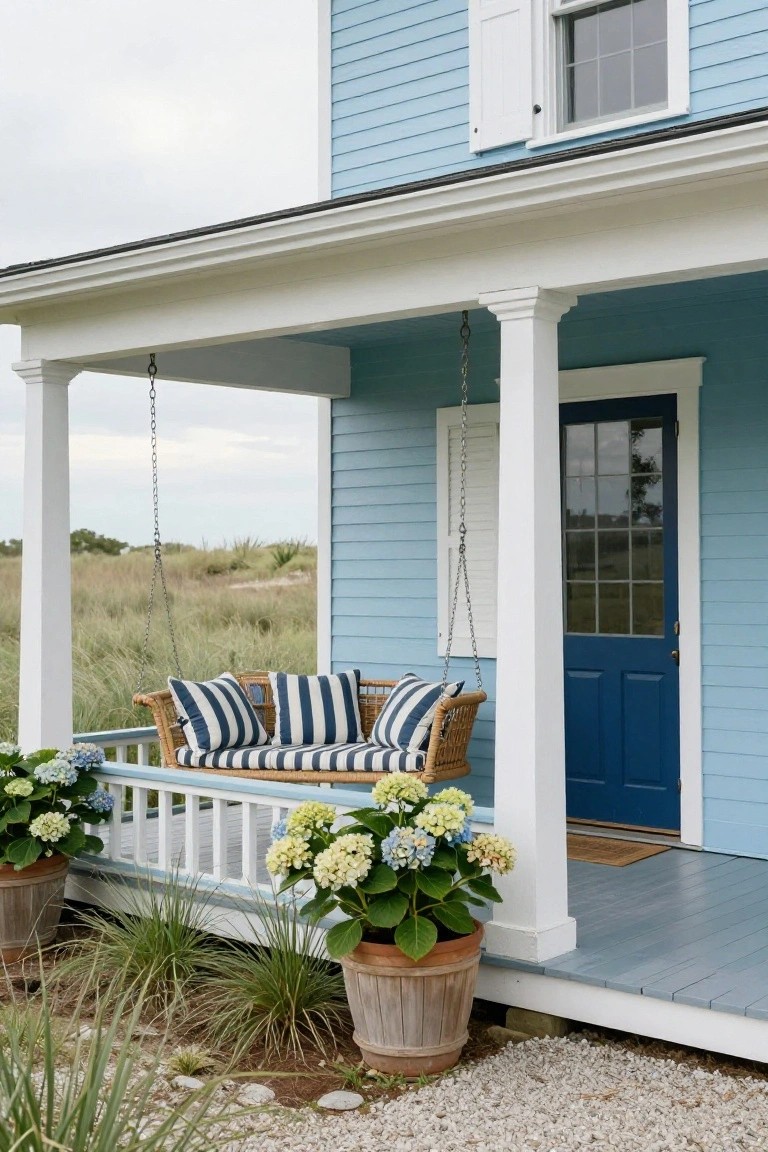 Light blue colonial house exterior with white trim and columns on a covered porch, hanging wicker swing with navy and white striped cushions and pillows, two large pots of hydrangeas, dark blue front door with glass panels, and dune grasses with gravel ground cover.