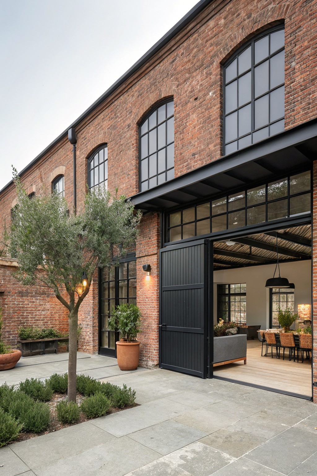 Red brick building exterior with large open black bi-fold doors revealing an interior space with wooden floors, sofa, dining table, and plants; olive tree, potted plants, and stone paving in front.