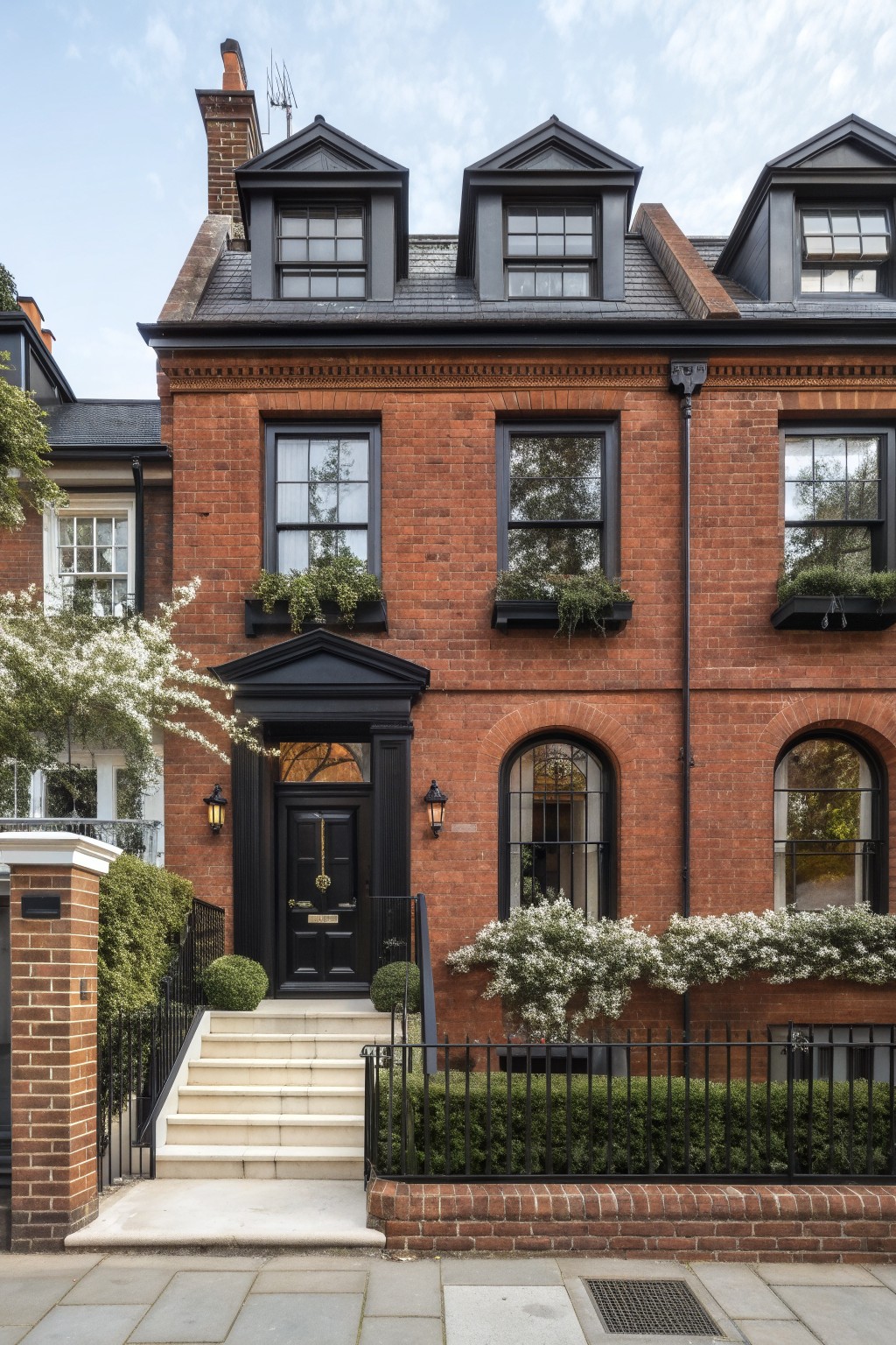 Three-story red brick townhouse with black trim on dormers, window frames, arched windows, and front door, stone steps with black railings, topiary spheres, and low hedges in front on a paved sidewalk.