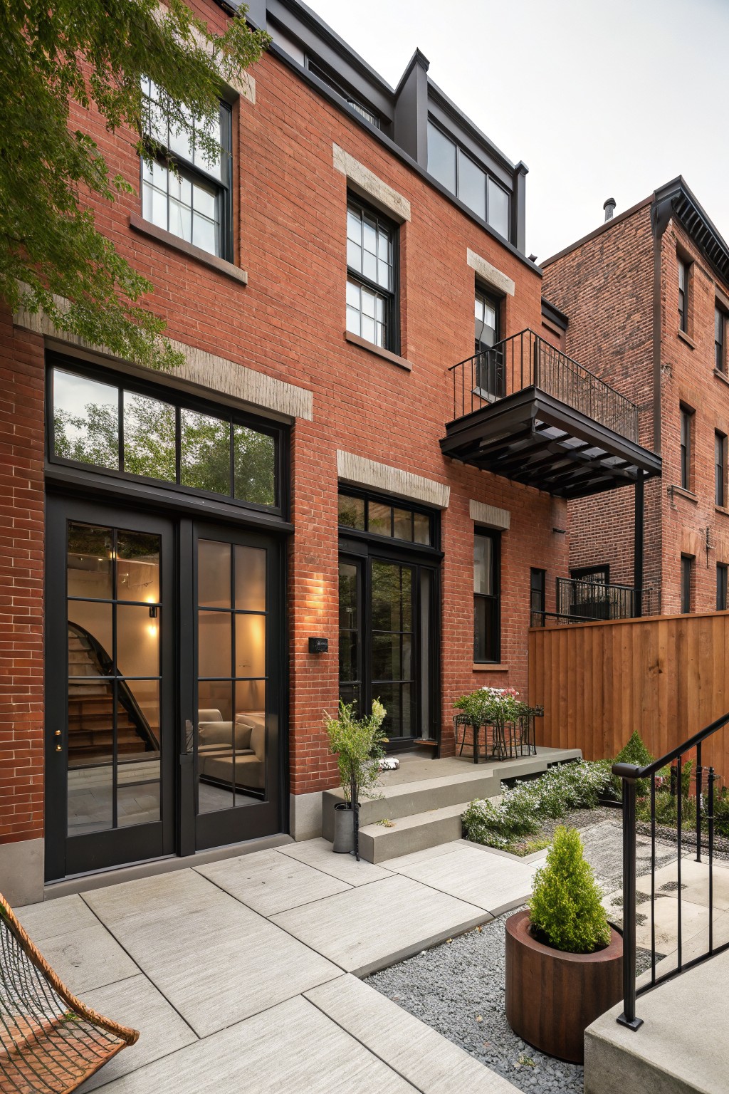 Rear view of a multi-story red brick townhouse with black-framed large glass doors, dark window trim, a metal balcony above, and a concrete patio area with potted plants, gravel, and wooden fencing.