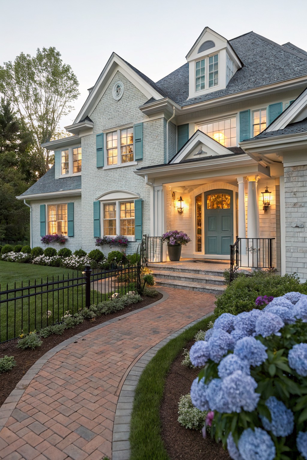 Two-story house with light gray painted brick exterior, teal shutters on windows, teal front door in arched entry porch with columns, black iron fence, brick pathway edged with blue hydrangeas and lawn, trees in background at dusk.