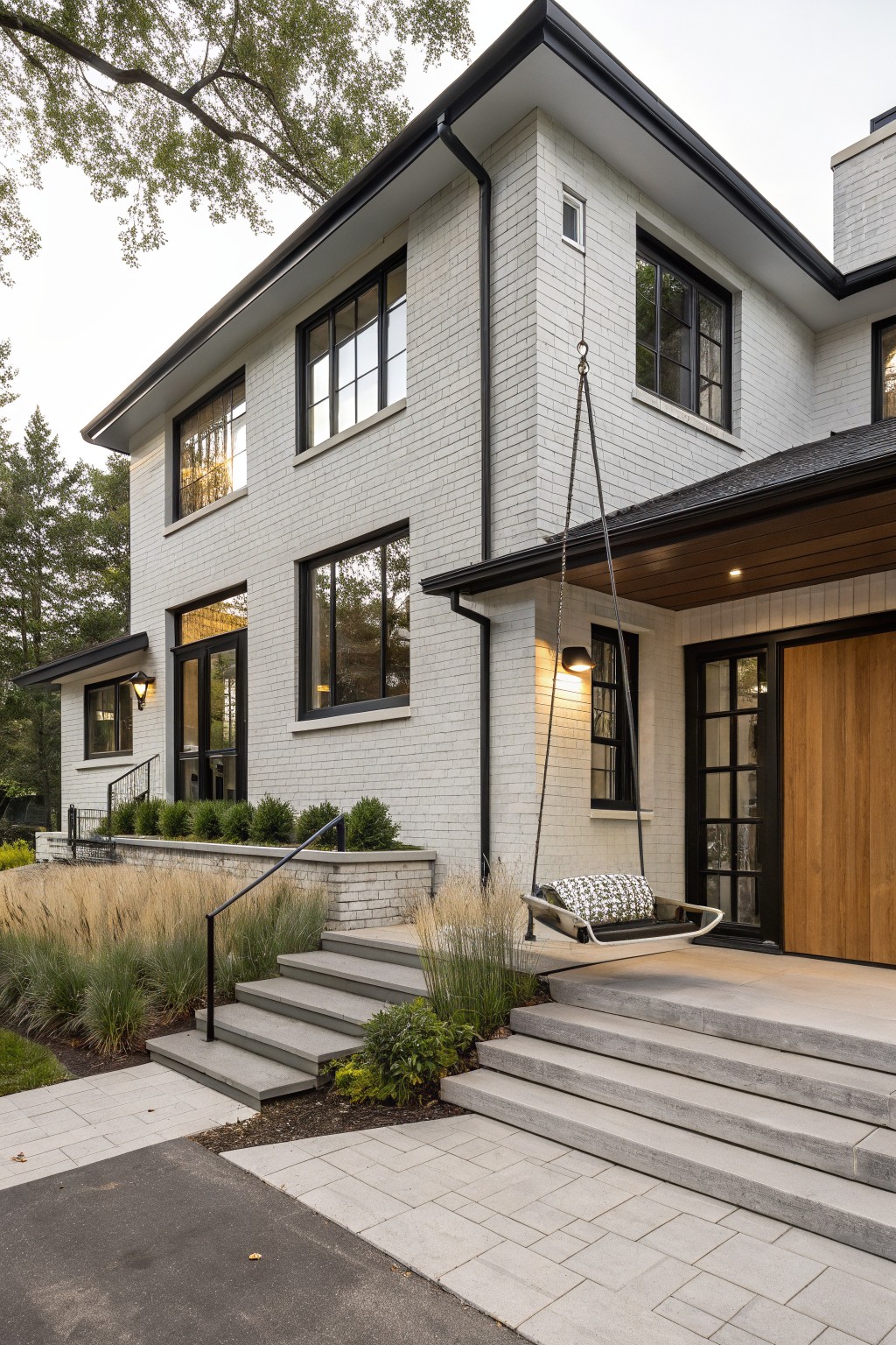 Two-story house exterior featuring white painted brick walls, black-framed windows and doors, a wooden front door with glass panels, a hanging porch swing, concrete steps, and ornamental grasses in the landscaping.