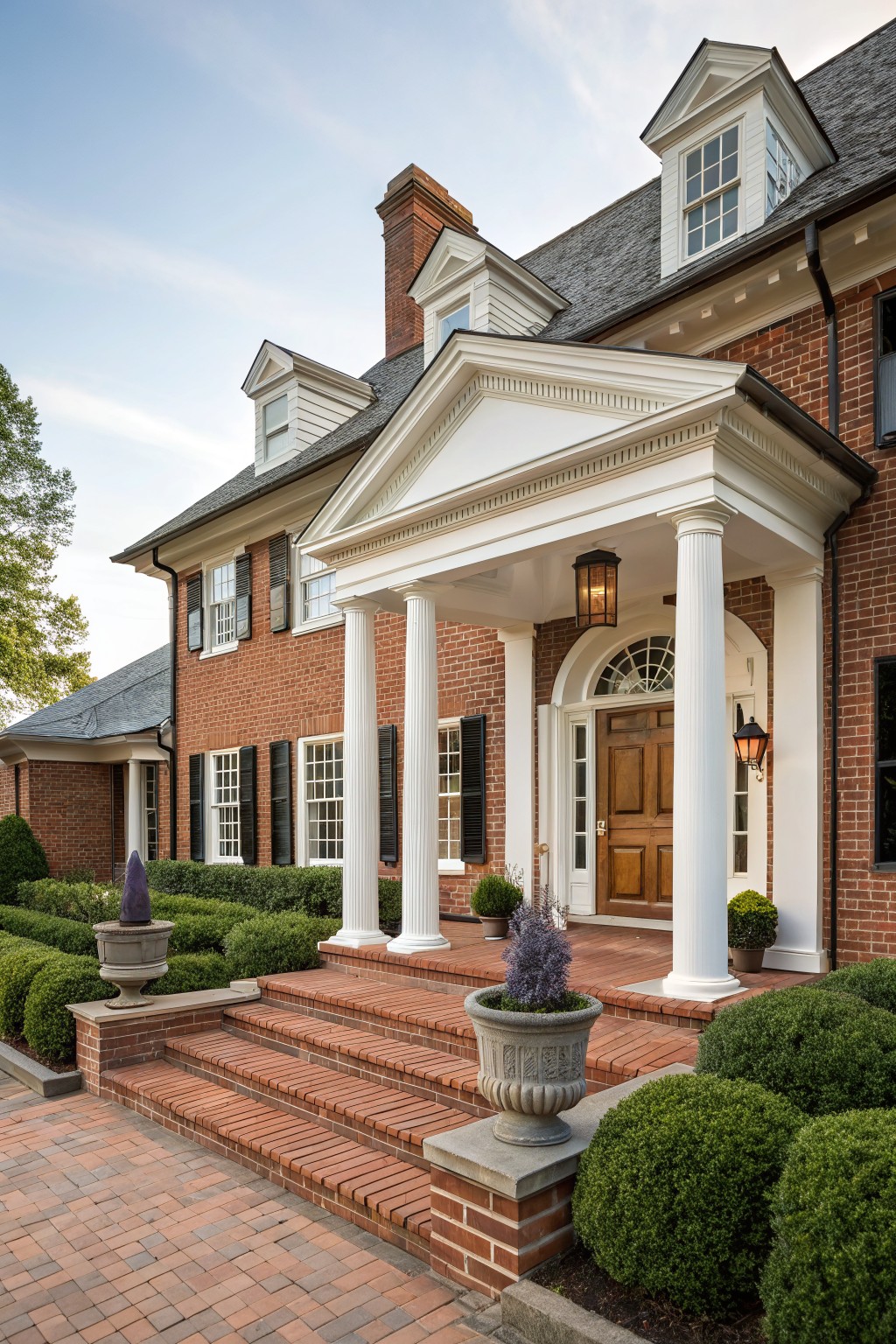 Red brick house exterior with white-columned portico, wooden front door, brick steps, lanterns, potted plants, and boxwood shrubs.