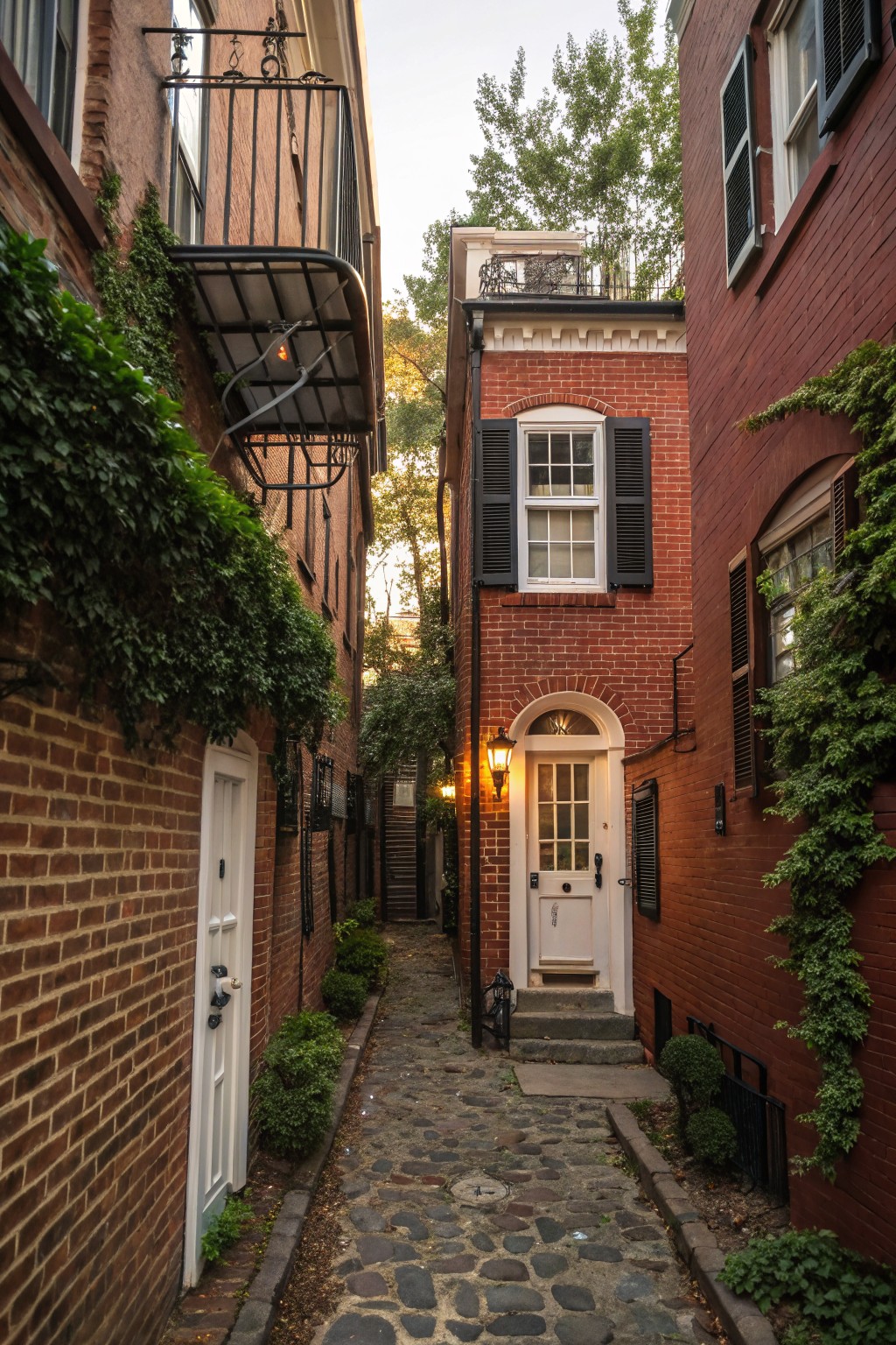 Narrow cobblestone alley flanked by red brick townhouse walls with white arched doors, ivy climbing the bricks, black lanterns, balconies, and shrubs.
