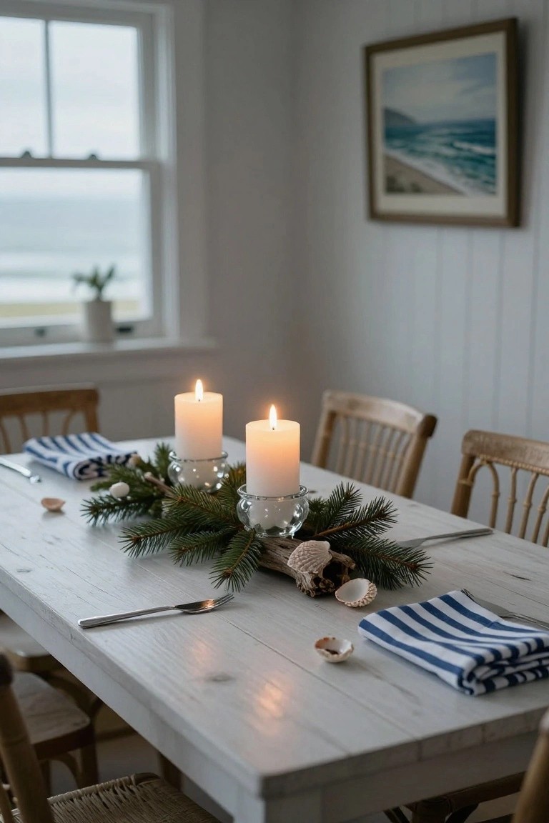 White wooden dining table with a centerpiece of fir branches, seashells, driftwood, and two lit white candles in glass holders, flanked by blue and white striped napkins, forks, and rattan chairs in a white paneled room with a window view of the ocean.