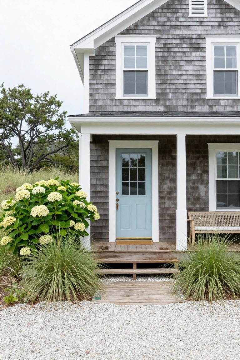 Gray shingled house with white trim, blue front door on a covered porch, large white hydrangea bush to the left, ornamental grasses, and gravel path in front.