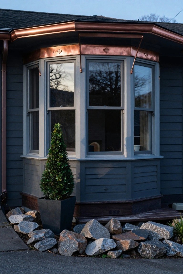 Dark gray shingled house exterior with copper gutters and trim around a multi-pane bay window, flanked by a potted topiary tree and rock garden bed.