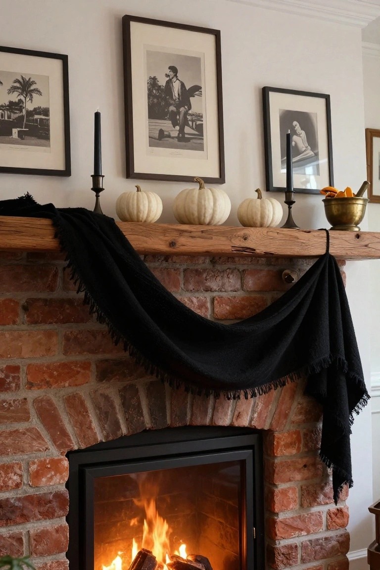 Rustic wooden mantel over a brick fireplace with burning fire inside, draped in fringed black fabric, decorated with white pumpkins, black taper candles in brass holders, a brass bowl of orange slices, and black-and-white framed photos on the wall.