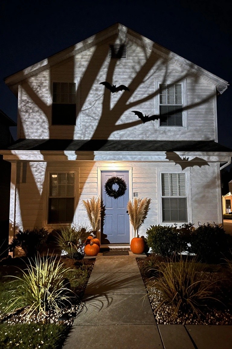 Nighttime view of a two-story white clapboard house with projected black bat shadows on the siding, blue front door with black wreath, pampas grass and orange pumpkins in front, and a concrete walkway.