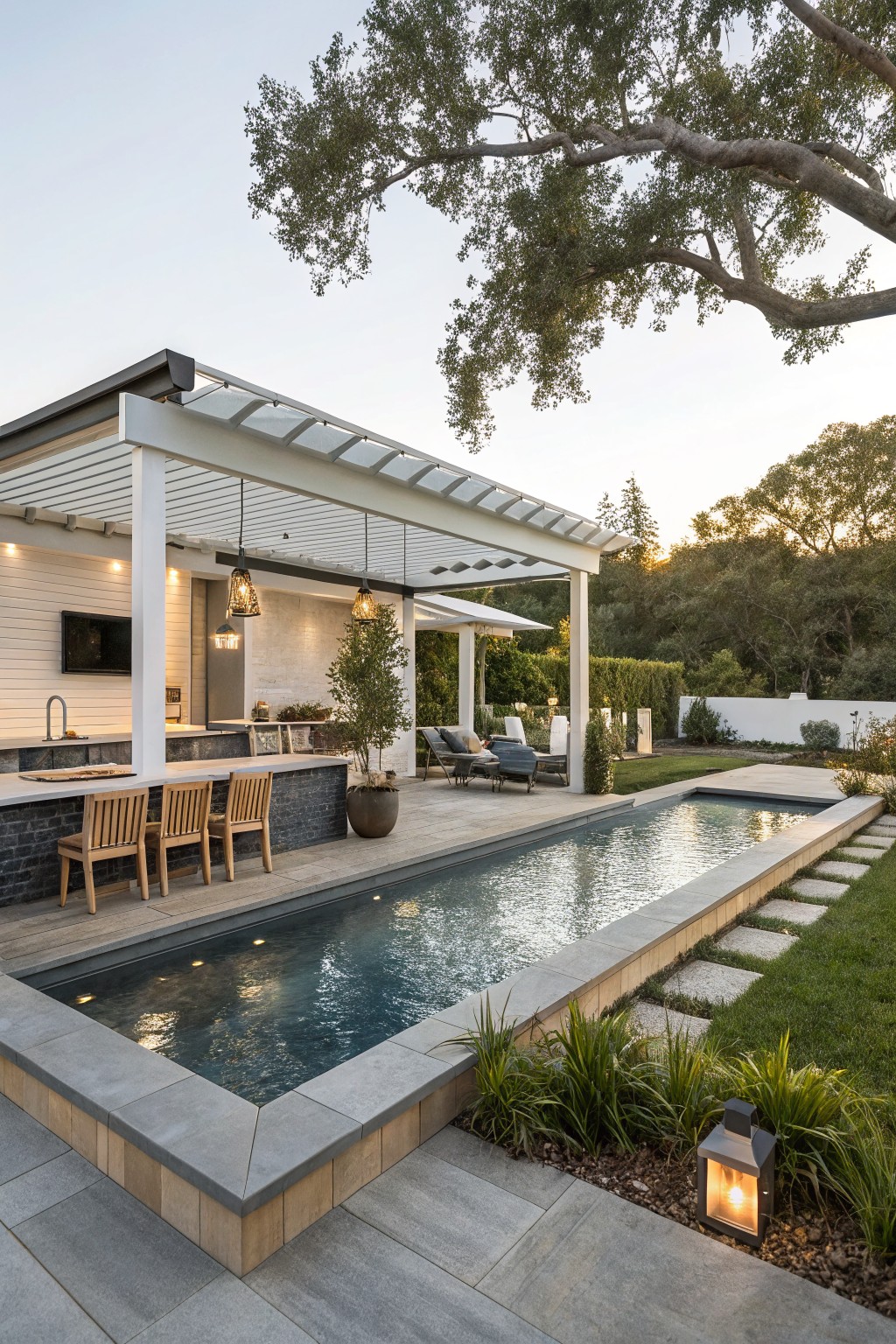 White outdoor bar with teak stools and sink under a slatted pergola, next to a narrow rectangular pool with underwater lights, lounge chairs nearby, and landscaping in a backyard setting.