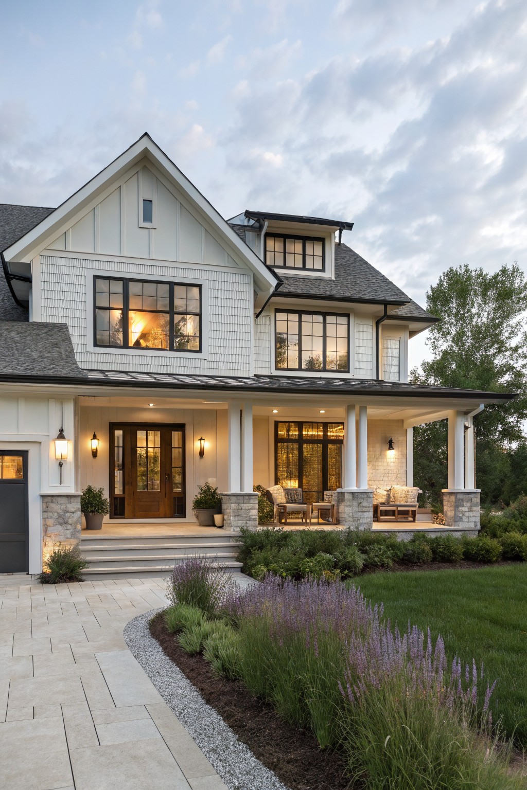 Two-story modern farmhouse exterior featuring white board-and-batten siding, multiple black-framed grid windows, covered front porch supported by white columns on stone bases, lanterns, seating furniture, double front doors, attached garage, and curved stone pathway edged with lavender plants and grasses.