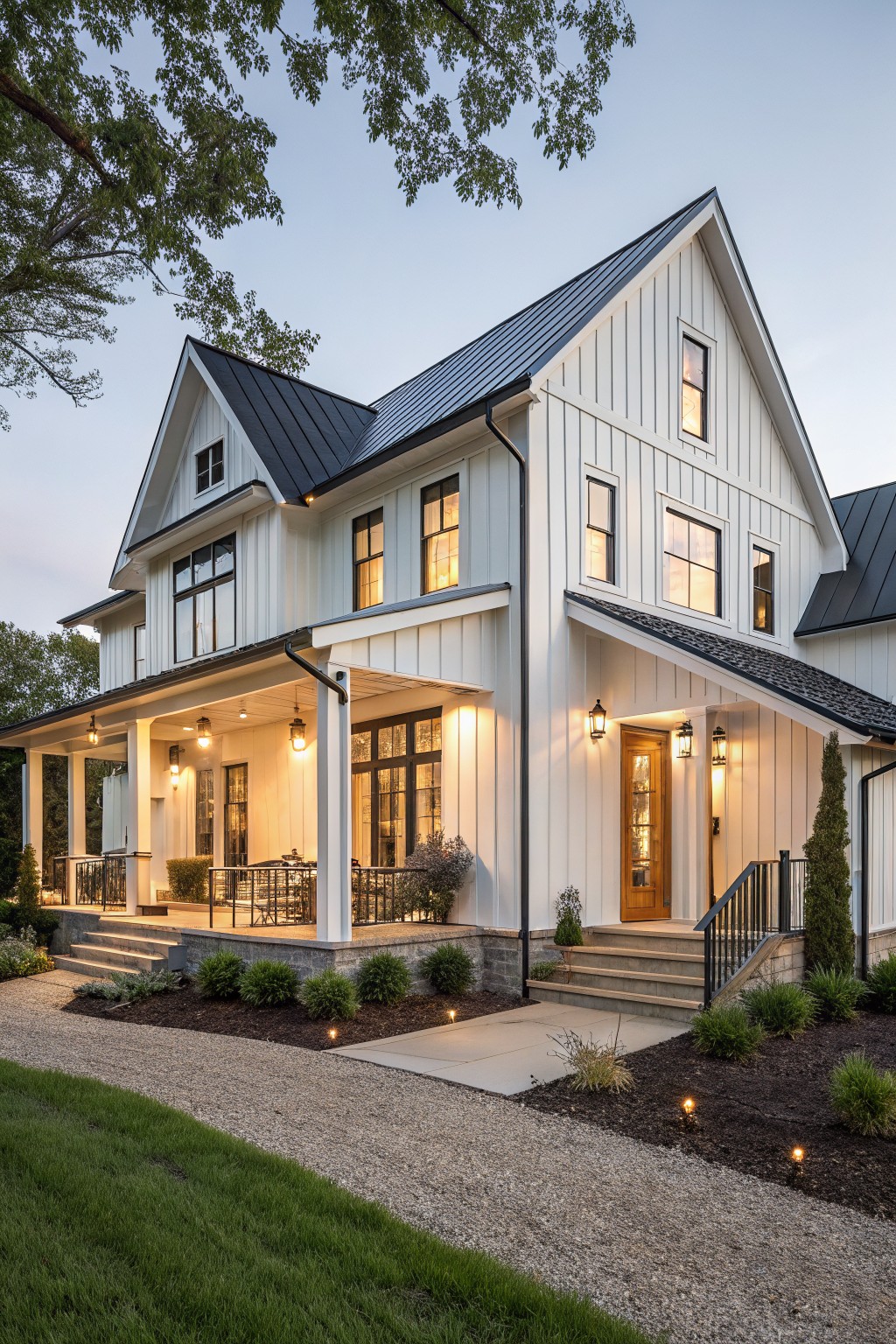 Two-story white board-and-batten farmhouse with black standing-seam metal roof, wrap-around porch, lanterns, and front landscaping at dusk.