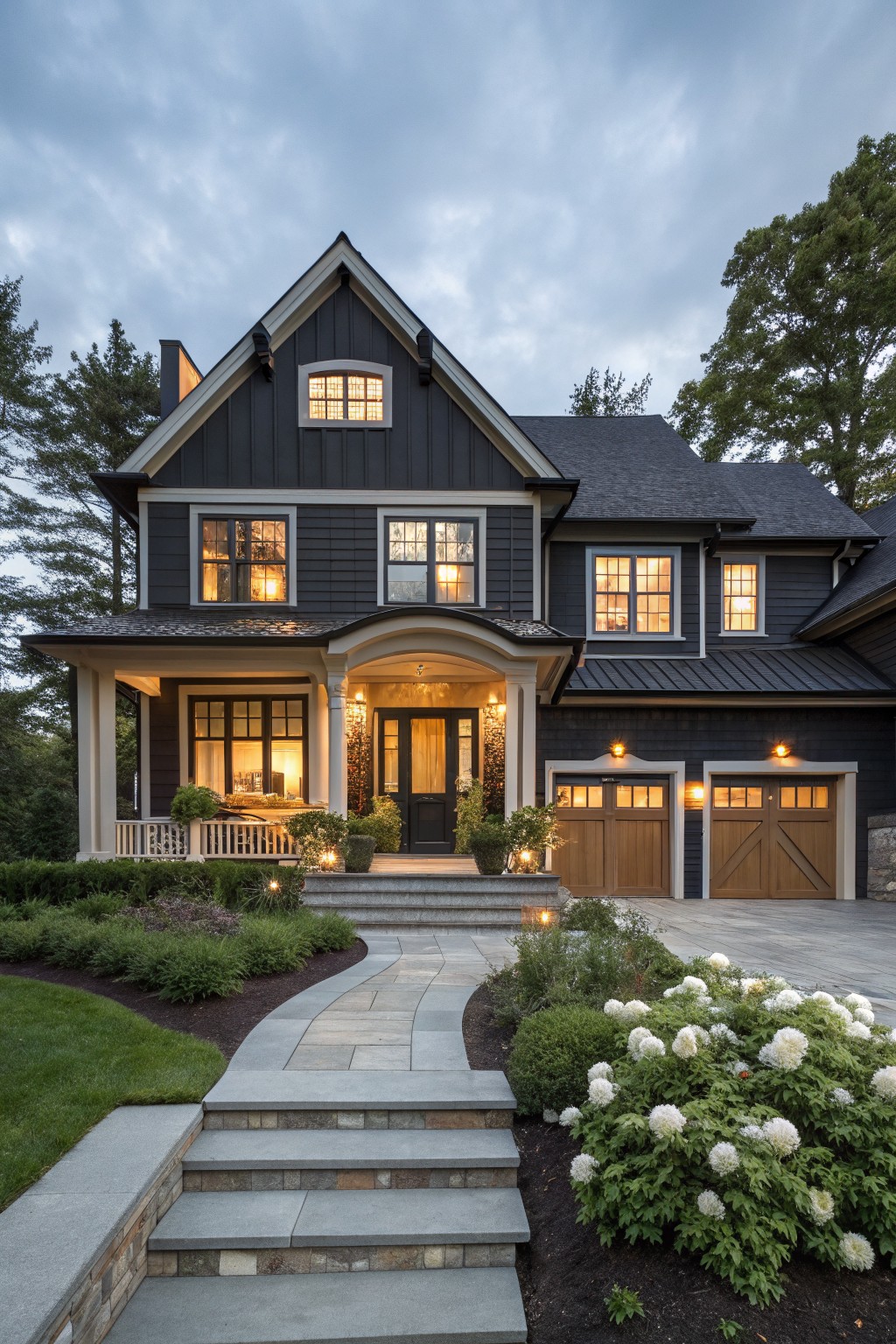 Two-story modern farmhouse exterior with dark gray shake siding, white trim, gabled roof, arched porch entry, wood garage doors, stone steps and pathway, and hydrangea plantings at dusk with interior lights glowing.
