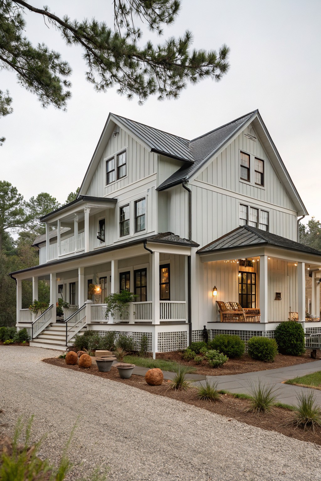 Two-story white board-and-batten farmhouse exterior with black metal roof, wraparound porch, black-framed windows, wooden stairs, potted plants, gravel driveway, and low landscaping with boulders.