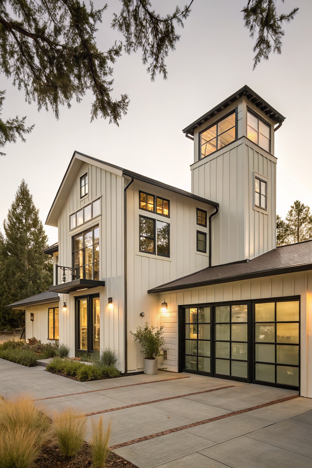 Two-story modern farmhouse exterior featuring white board-and-batten siding, a tall central tower with windows, attached garage with glass doors, pathway, and surrounding trees and grasses at dusk.