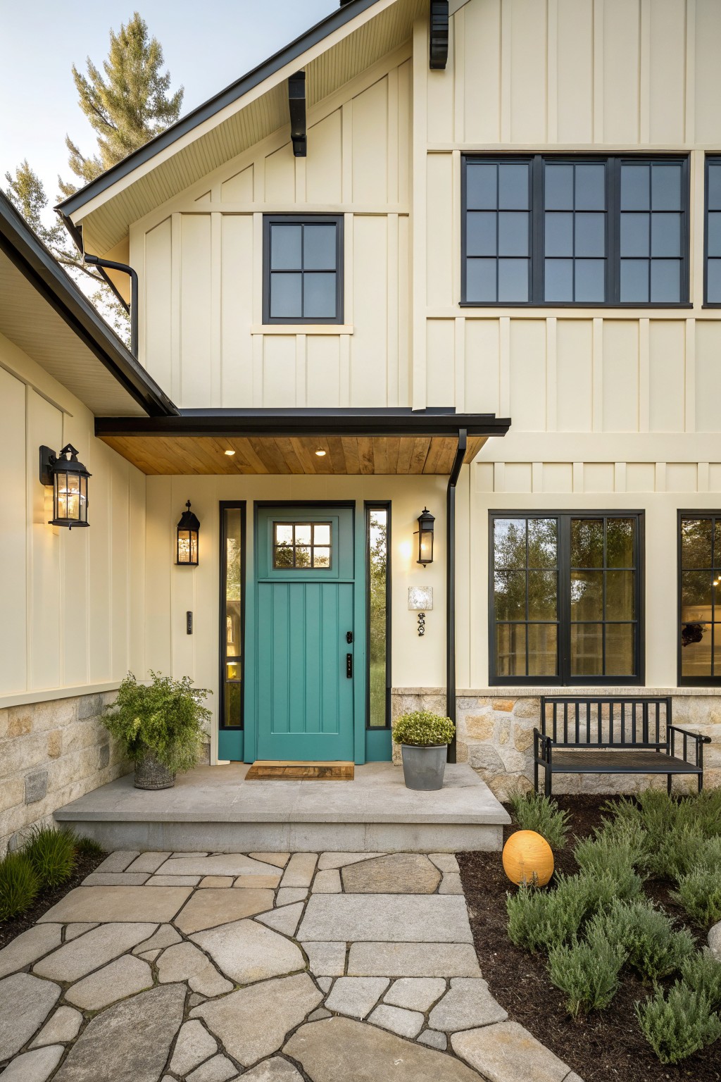 Front exterior of a modern farmhouse with creamy beige vertical siding, teal paneled front door, black-framed windows, wooden porch overhang, stone foundation, black metal bench, potted plants, and flagstone pathway.