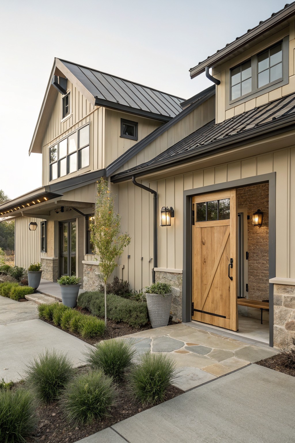 Beige board-and-batten sided modern farmhouse exterior with black standing-seam metal roof, open wooden barn door entry, lanterns, potted plants, and low landscaping along a stone path.