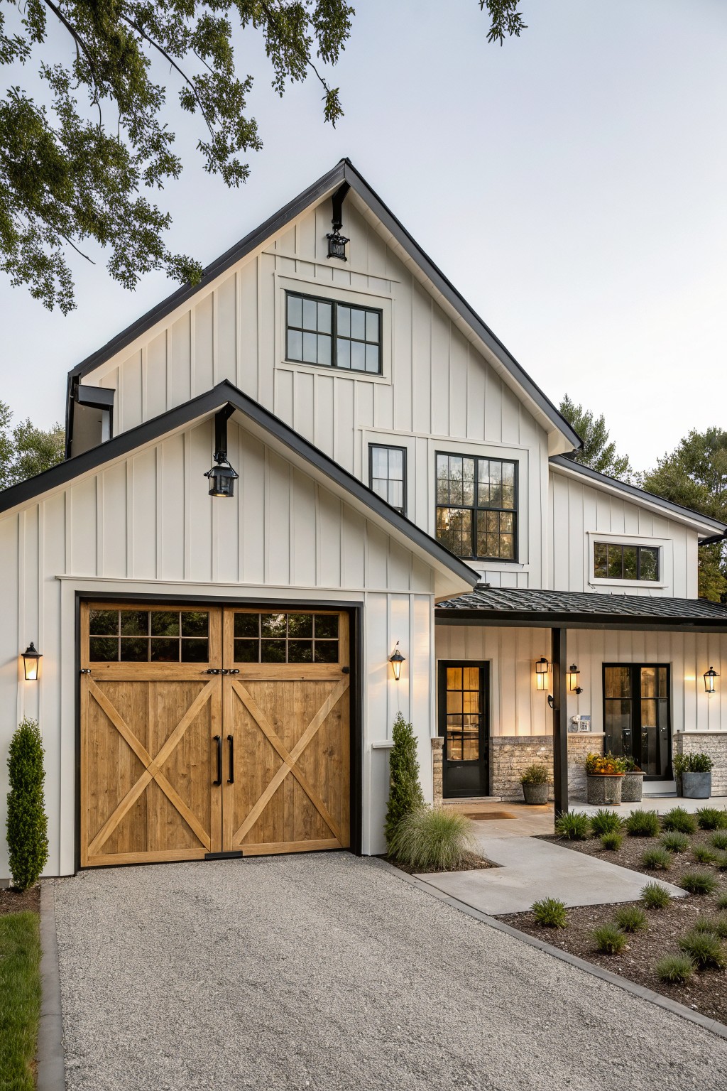 Two-story modern farmhouse house exterior with white board-and-batten siding, black metal gable roof, black-framed windows, wooden garage doors, covered front porch, gravel driveway, and low plantings.