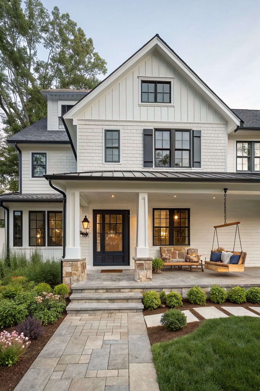 Two-story modern farmhouse with white shiplap siding, black window frames and door, dark gray roof, covered porch with hanging swing and stone pillars, surrounded by landscaping and a stone pathway.