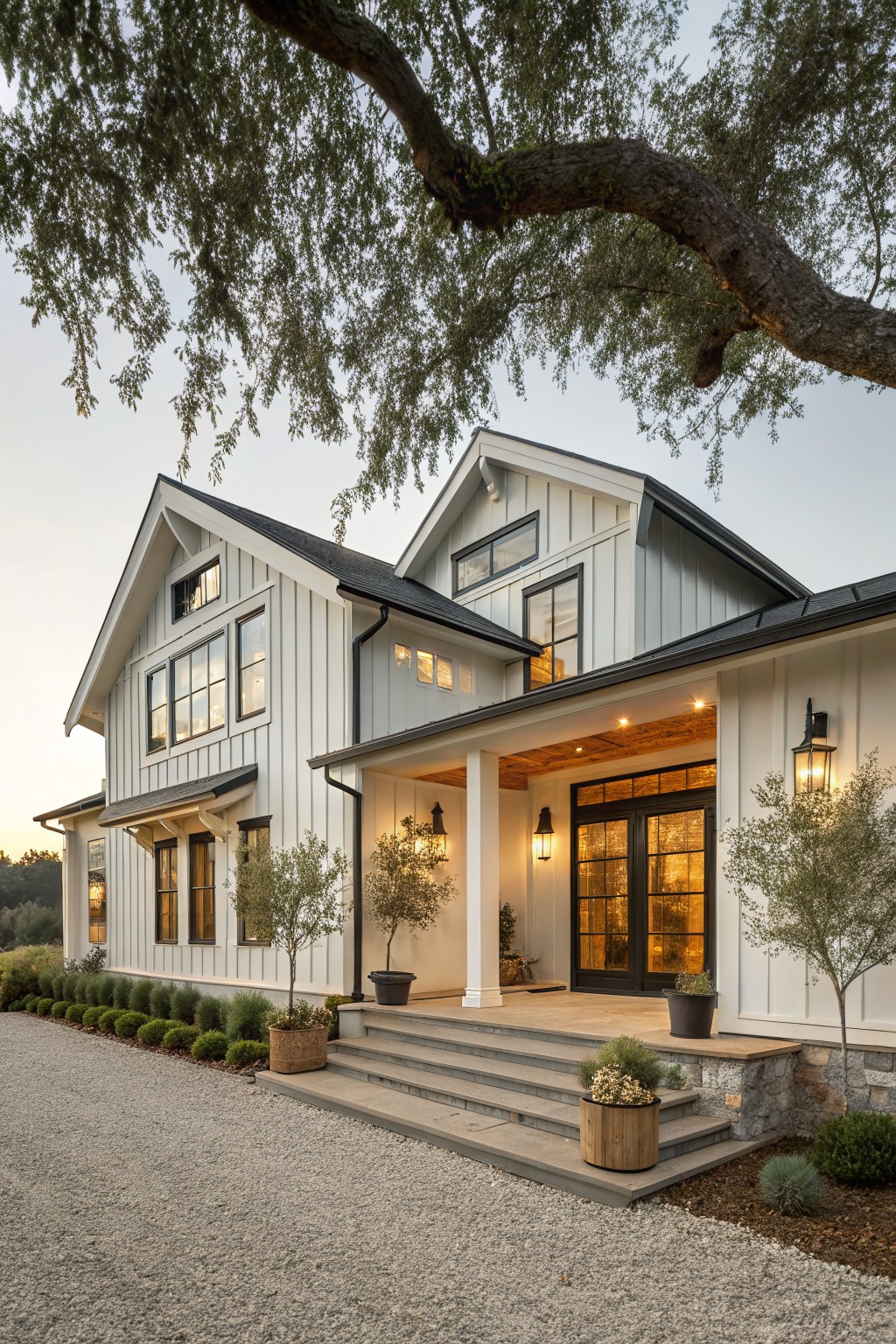 White board-and-batten sided modern farmhouse with dark metal gable roof, black trim and windows, covered front porch with wood beams, stone steps, olive trees flanking the entry, gravel driveway, and low shrubs.