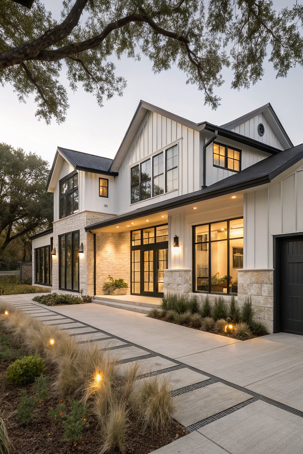 Two-story modern farmhouse exterior with white board-and-batten siding, dark metal gable roof, black garage door, stone entry accents, large black-framed windows, concrete pathway, and ornamental grasses under live oak trees at dusk.