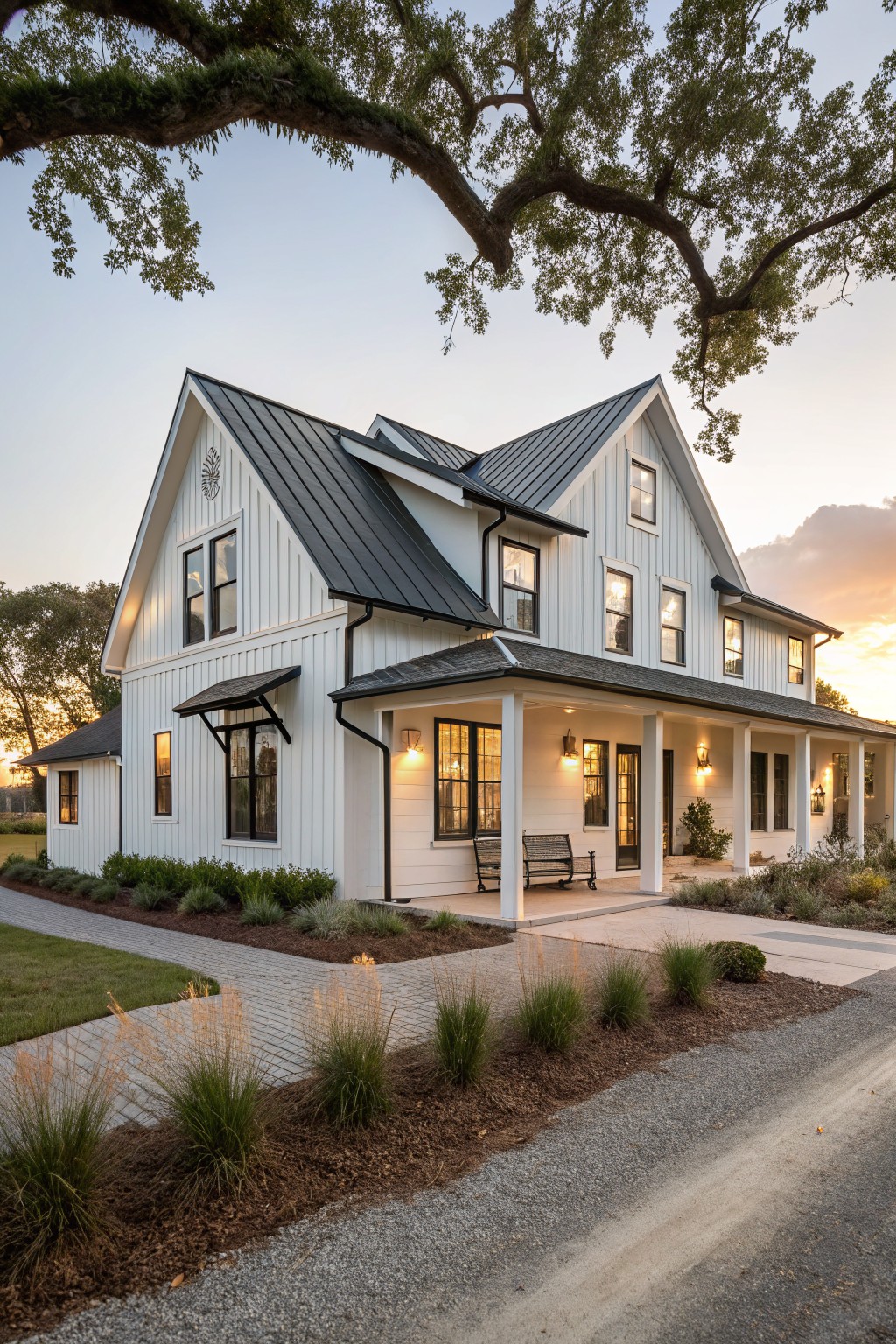 Side view of a two-story white board-and-batten farmhouse with black standing-seam metal gable roof, wrap-around porch with columns and bench, downspouts, flanked by grasses and shrubs under a large oak tree at sunset.